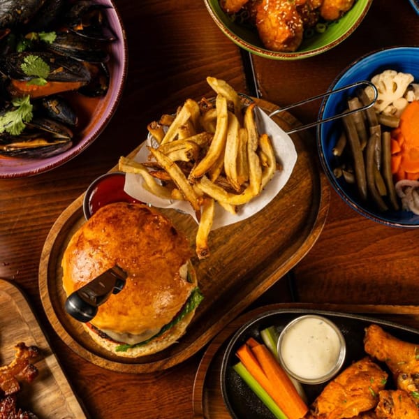 Overhead view of a burger with fries, mussels, chicken wings, and pickled vegetables on a wooden table.