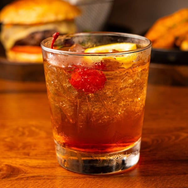 A Peggy's cocktail with a cherry and lemon slice, with a burger in the background.