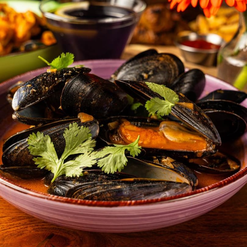 Steamed mussels in a rich red broth, garnished with fresh cilantro, served in a purple bowl.