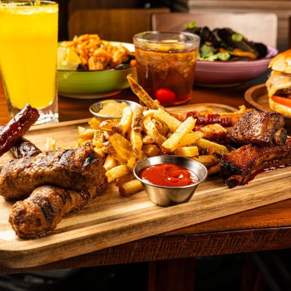 Grilled European sausages, ribs, and fries with ketchup on a wooden board, with drinks and other dishes.