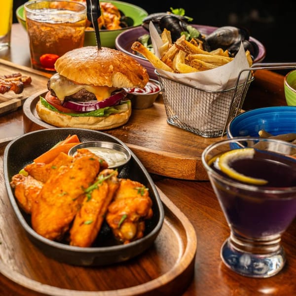 A Peggy's burger, fries, chicken wings, mussels, and drinks on a wooden table.