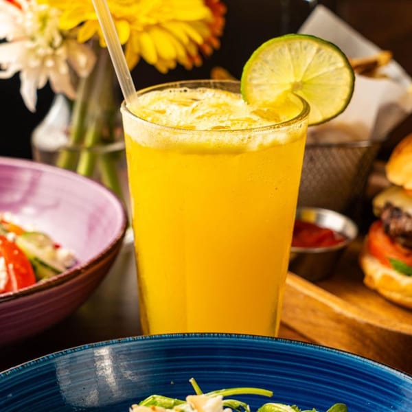 A vibrant orange drink with a lime slice and straw, next to a salad and burgers.